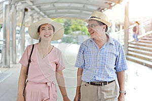 Asian traveler couple husband and wife in pink and blue shirt with white nice hat and luggage walking in downtown with happy