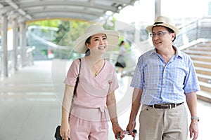 Asian traveler couple husband and wife in pink and blue shirt with white nice hat and luggage walking in downtown with happy