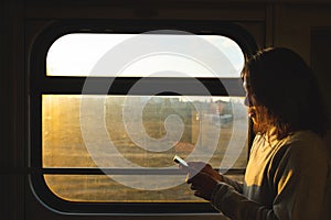 Asian traveler woman looks at smart mobile phone next to the window of a moving train. Sunset sky and the silhouette girl against