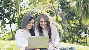 Asian two women talking using laptop sitting on green meadow at park. Friendship girls chat and talk look at laptop planning do