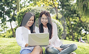 Asian two women talking using laptop sitting on green meadow at park. Friendship girls chat and talk look at laptop planning do