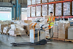 Asian warehouse worker stand and drive folklift in front of stack of product carton in workplace also look at camera