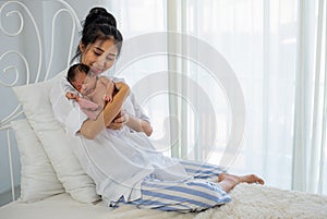 Asian white shirt mother holds her little sleeping newborn baby on her chest and sit on white bed in front of glass windows with