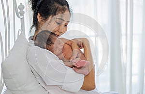 Asian white shirt mother holds her little sleeping newborn baby on her chest and sit on white bed in front of glass windows with