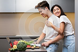 Asian Wife Hugging Husband While He Cooking Dinner In Kitchen