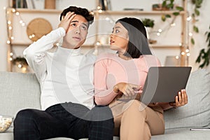 Asian Wife Shopping Online With Shocked Husband Sitting At Home