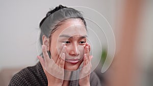 An Asian woman applying face cream to nourish her skin in front of a large mirror after washing and styling at home.