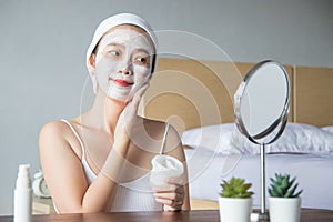 Asian woman applying moisturizer face scrub peeling white clay mask on skin, looking in mirror with smiley face using cream