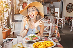 Asian woman in big hat having meal with greek salad Horiatiki in restaurant. Greece cuisine concept