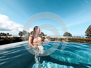 Asian woman in bikini in infinity swimming pool edge. luxury vacation near the sea.
