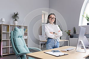 Asian woman businesswoman in the office at the desk. Crossed her arms, looks at the camera, smiles