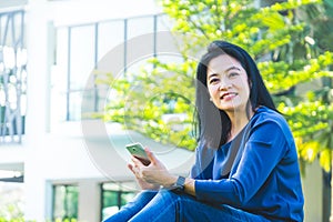 Asian woman chatting on mobile phone and smiling at screen sitting on grass outside home