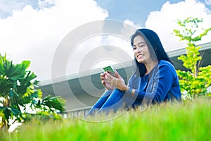 Asian woman chatting on mobile phone and smiling at screen sitting on grass outside home,Low angle view