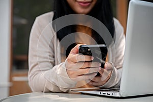 An Asian woman checking messages on her smartphone while working on her laptop at a cafe