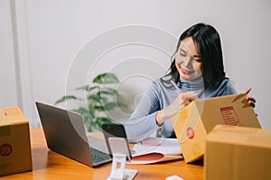 Asian woman checking sales on smartphone and working on laptop computer from home on table with parcel, e-commerce business,