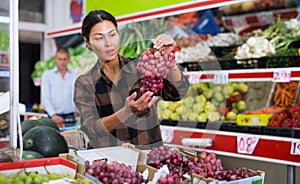 Asian woman choosing ripe grapes in greengrocery store
