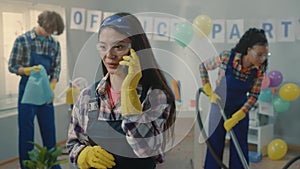 Asian woman cleaner talking on a cell phone and smiling. A European man puts garbage in a trash bag while an African