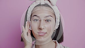 asian woman cleans her face with a cotton pad in the bathroom in pink pajamas and a headband.