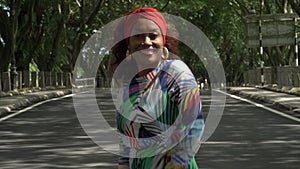 Asian woman in a colored stylish dress with turban walks and dance along the asphalt road under shady trees in the park.