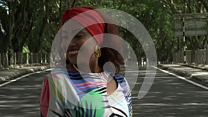 Asian woman in a colored stylish dress with turban walks and dance along the asphalt road under shady trees in the park.