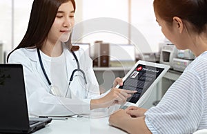 Asian woman doctor holding tablet with patient medical record on screen while consulting patient in medical consultation room.