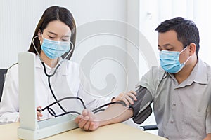 Asian woman doctor uses a blood pressure meter with a man patient to check his health at hospital. They wear a medical face mask