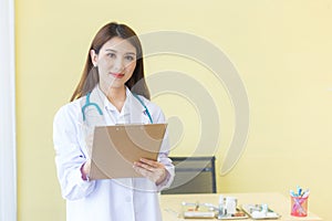 Asian woman doctor who wears medical coat and stethoscope is writing data in document of clip broad in hospital