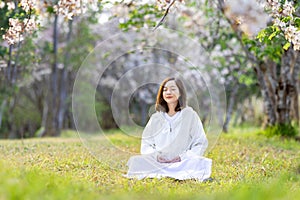 Asian woman is doing meditation under flower tree during cherry blossoming season for inner peace, mindfulness and zen practice