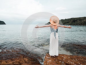 Asian woman in dress and straw hat looking away to the turquoise calm sea water. female open arms on the rock at tropical beach in