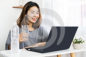 Asian woman drinking fresh water at work place sitting at office hand typing on laptop and holding glass of water