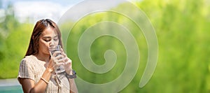 Asian Woman drinks water from tall glass of water at outdoor tree bokeh green for banner background