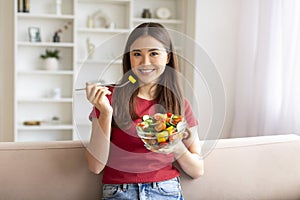 Asian woman eating vegetable salad while sitting on couch in living room