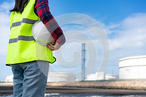 Asian woman engineer holding a white safety helmet with oil refinery tank plant factory in background with blue sky