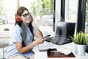 Asian woman with eyeglass using a mobile phone and headphones with a laptop and a notebook with a cup of coffee on the table