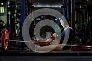 Asian woman is facing overtraining syndrome after weight training workout inside gym with dark background for exercising and