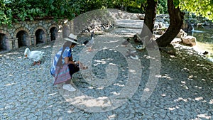 Asian woman with fedora and sunglasses kneeling in front of ducks and geese and looking away from photographer