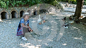 Asian woman with fedora and sunglasses kneeling in front of ducks and geese and looking at photographer