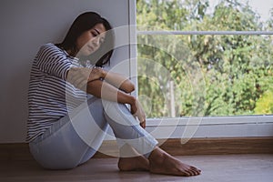 An Asian woman feeling sad and lonely sitting by the window in the house. Confusion, disappointment, and mental problems