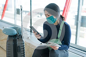 Asian woman flying in covid19 times - young tired and attractive Chinese girl in face mask waiting on airport checking mobile