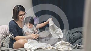 Asian woman folding daughter clothes on bed, Little baby sit on mom try to help mother, Living lifestyle family indoors concept