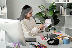 Asian woman freelance graphic designer working with color swatch samples and computer at desk in home office, young lady
