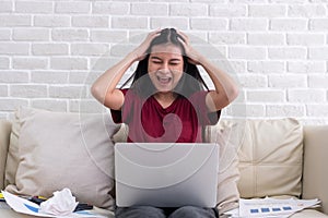 Asian woman freelancer shout with stress emotion while working with laptop and paperwork at sofa in living room in house.