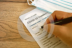 An Asian woman hand writing filling Covid-19 vaccine consent paper form with a black pen with medical mask on the table. Health ca