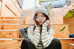 Asian woman in hat expressing sadness while sitting on stairs indoors