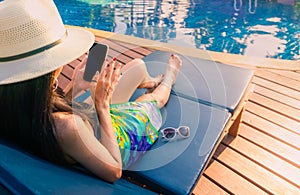 Asian woman with hat and swimsuit sit on sunbed at poolside and using smartphone on summer vacation by swimming pool. Girl