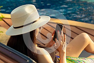 Asian woman with hat and swimsuit sitting in chair at poolside and using smartphone on summer vacation. Luxury life. Checking News