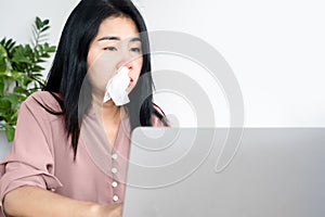 Asian woman having allergic rhinitis  because of cold weather and dust sitting at office desk with tissue in her nose