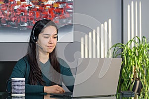 Asian woman having a business call in headphones at a desk, working on laptop in home office