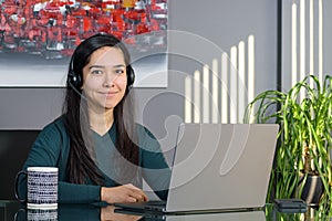 Asian woman having a business call in headphones at a desk, working on laptop in home office
