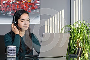 Asian woman having a business call in headphones at a desk, working on laptop in home office